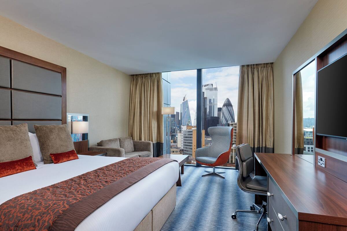 A hotel room with a large bed, a desk, a chair, and a view of the city skyline.
