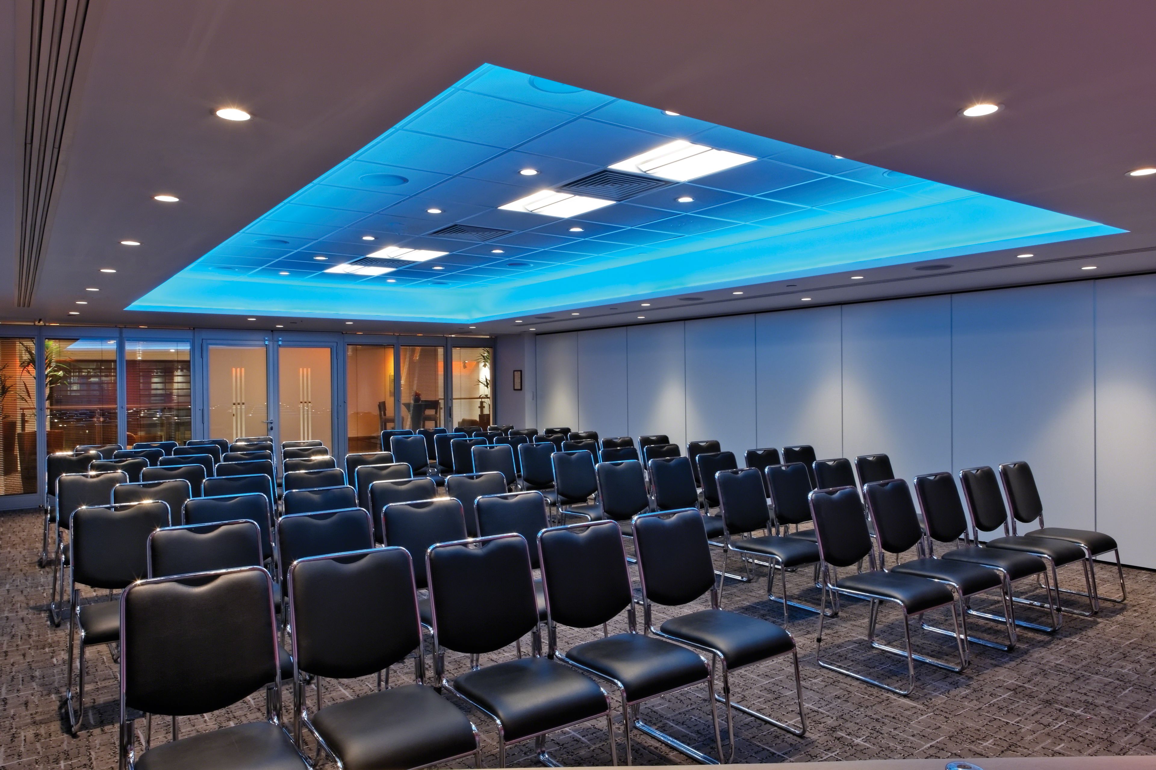 A conference room with rows of black chairs, a blue-lit ceiling, and large windows.