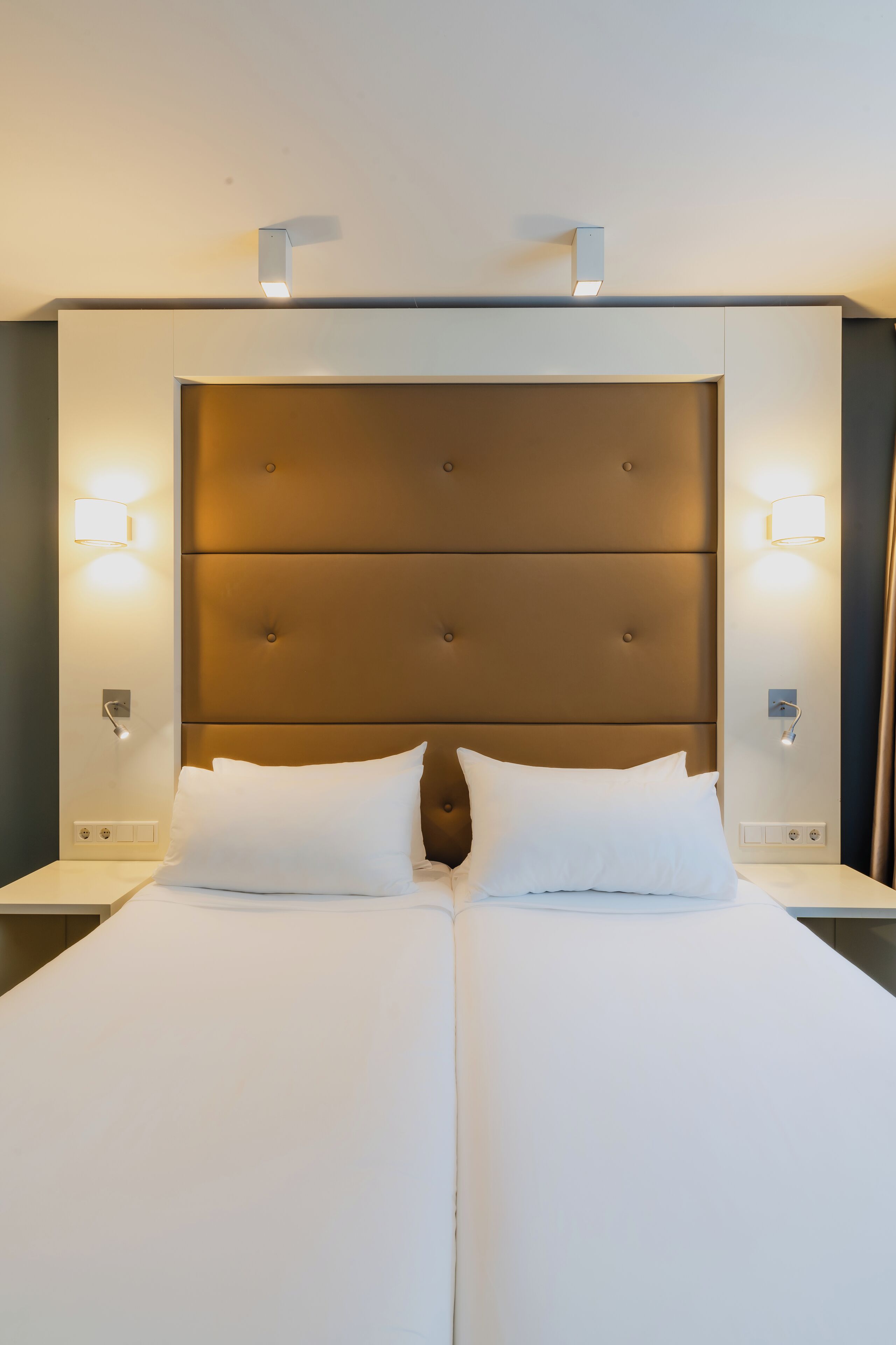 Un lit double avec une tête de lit capitonnée, deux tables de chevet et des lampes fixées au mur.