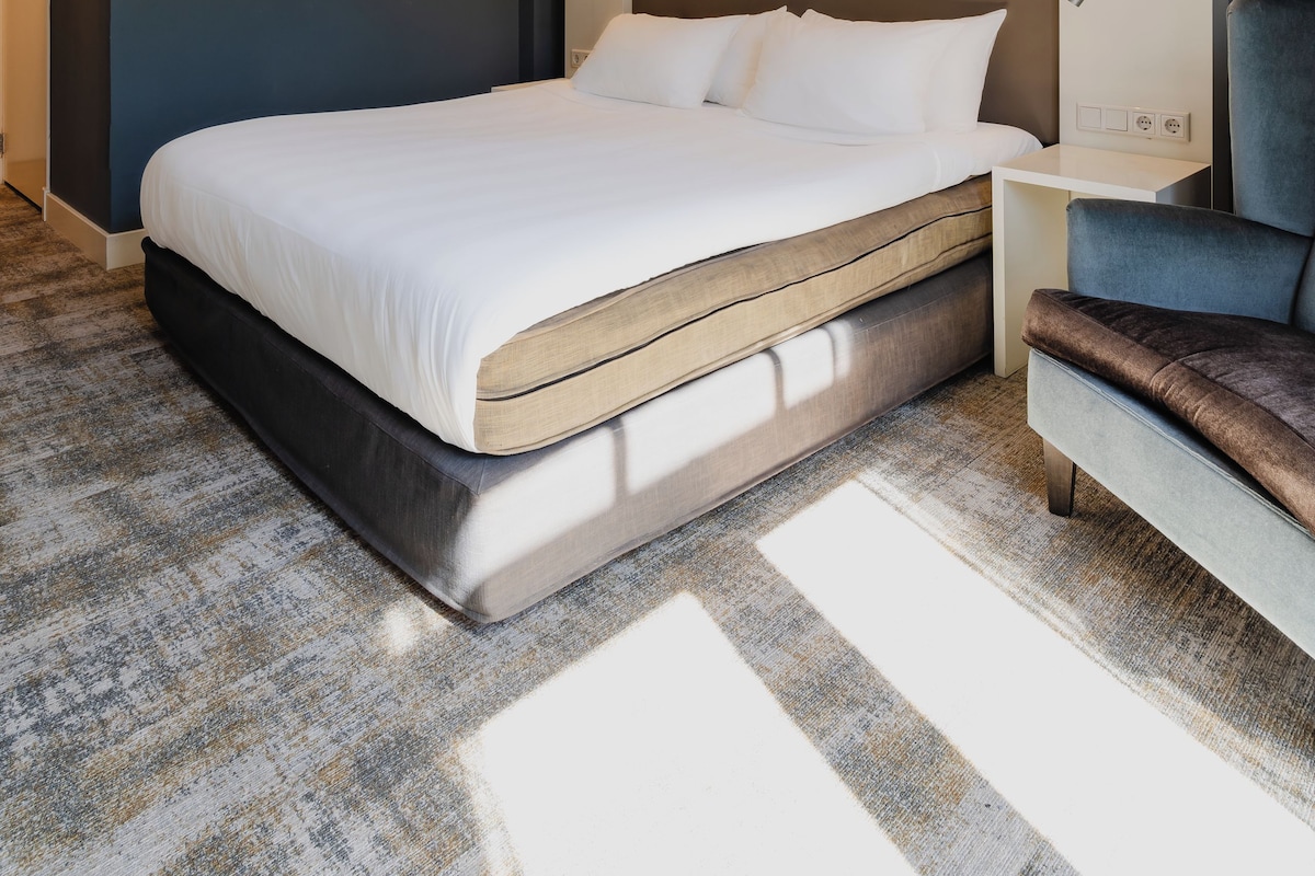 A modern hotel room with a large bed, a nightstand, a chair, and a wall-mounted light fixture.