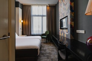 Ein Hotelzimmer mit Bett, Schreibtisch, Sessel, Fernseher und einem Fenster mit Vorhängen.