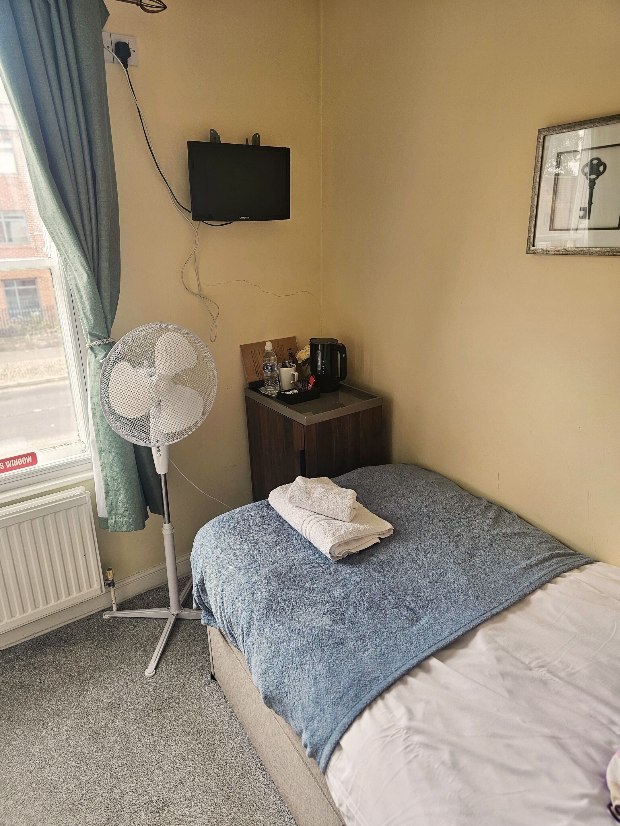 A hotel room with a bed, a standing fan, a coffee maker, and a TV on the wall.