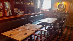 Interior de restaurante com mesas e cadeiras de madeira, área de bar e uma estante com livros.