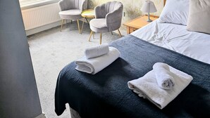 Quarto de hotel com uma cama, duas poltronas, uma mesa pequena, um abajur e uma janela com cortinas.