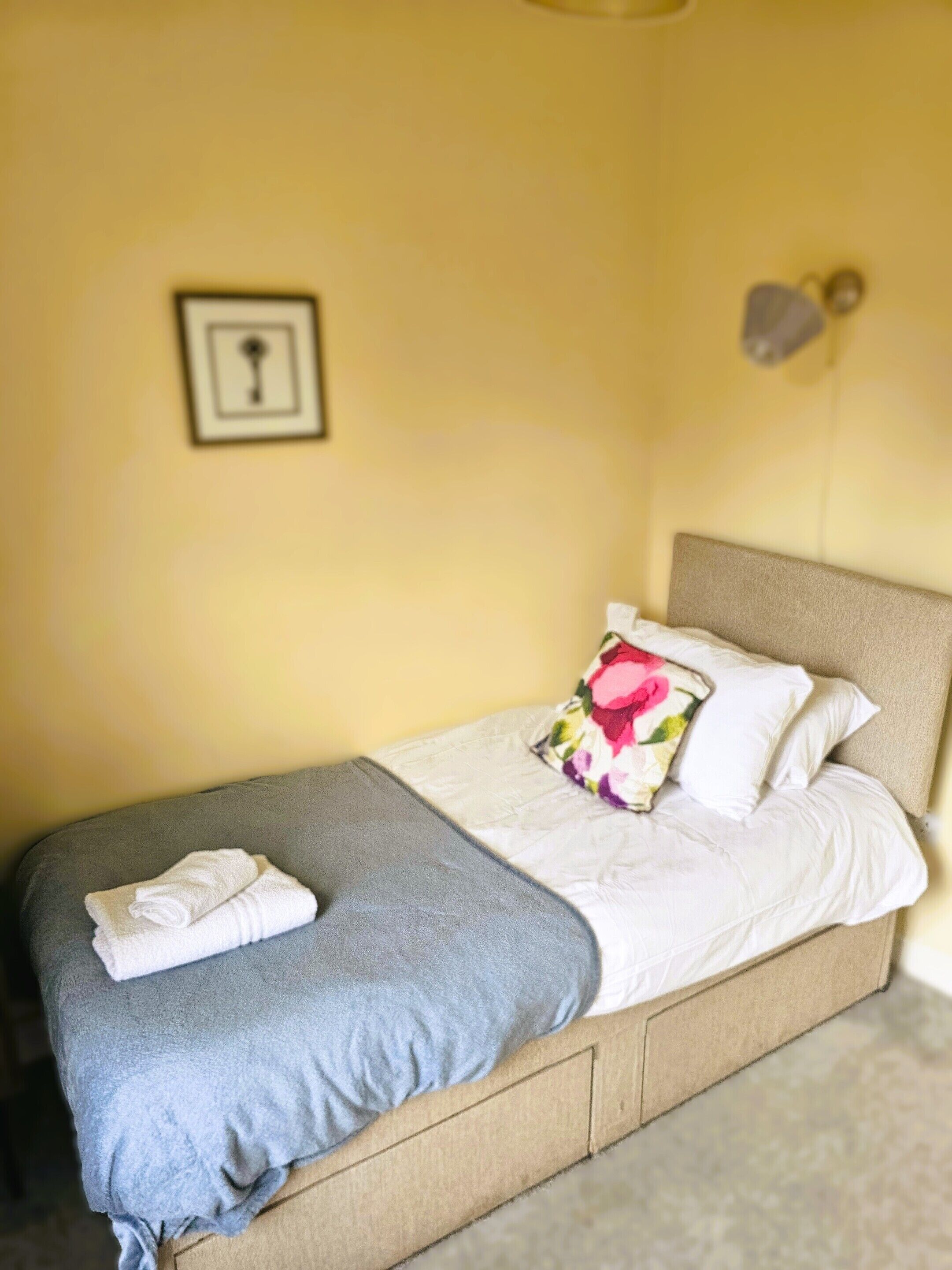 A neatly made bed with a grey blanket and a folded white towel, a floral pillow, and a headboard.