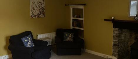 A living room with a fireplace, a black sofa, a black armchair, a bookshelf, and a floral painting on the wall.