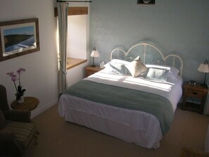 A bedroom with a large bed, a nightstand, a chair, a small table with a plant, and a window with curtains. - The Who'd Have Thought It Inn (Yelverton)