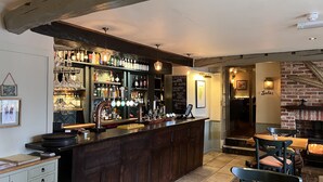 Een bar met een houten toonbank, flessenplanken en een krijtbordmenu met cocktails. Er is een zithoek met een open haard en een bord waarop 'Ladies' staat.