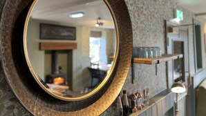 Een spiegel met gouden lijst weerspiegelt een kamer met bloemenbehang, een open haard en een plank met flessen.