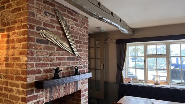 A room with a brick fireplace, a decorative feather, and a window with curtains. - The Forresters Arms Kilburn (York)