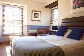 A bedroom with a large bed, a chair, and a desk. There are windows with blinds, a framed picture on the wall, and a radiator under the bed.