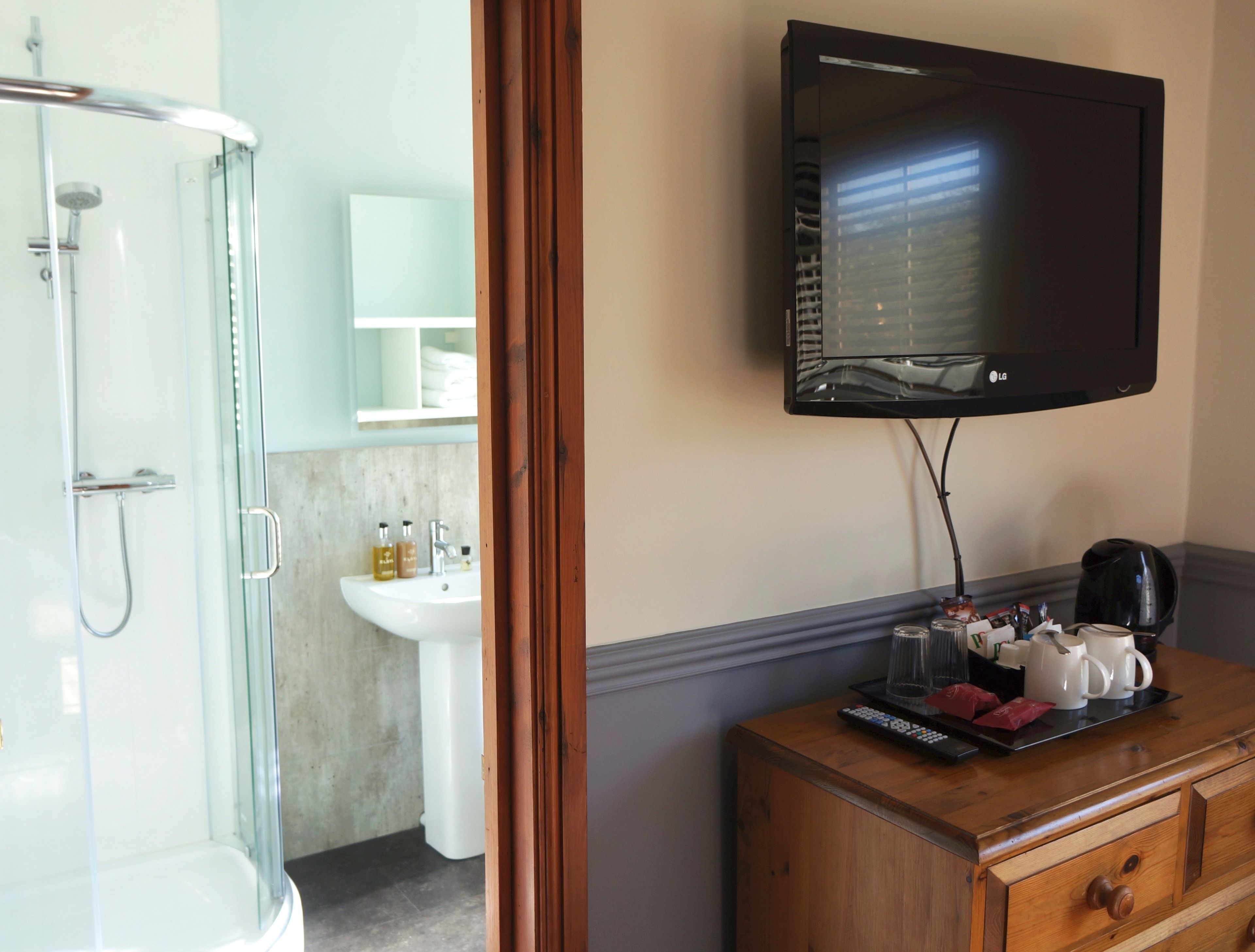 A bathroom with a glass shower enclosure, a sink, and a mirror. There is a television mounted on the wall, a wooden dresser, and a tray with toiletries.