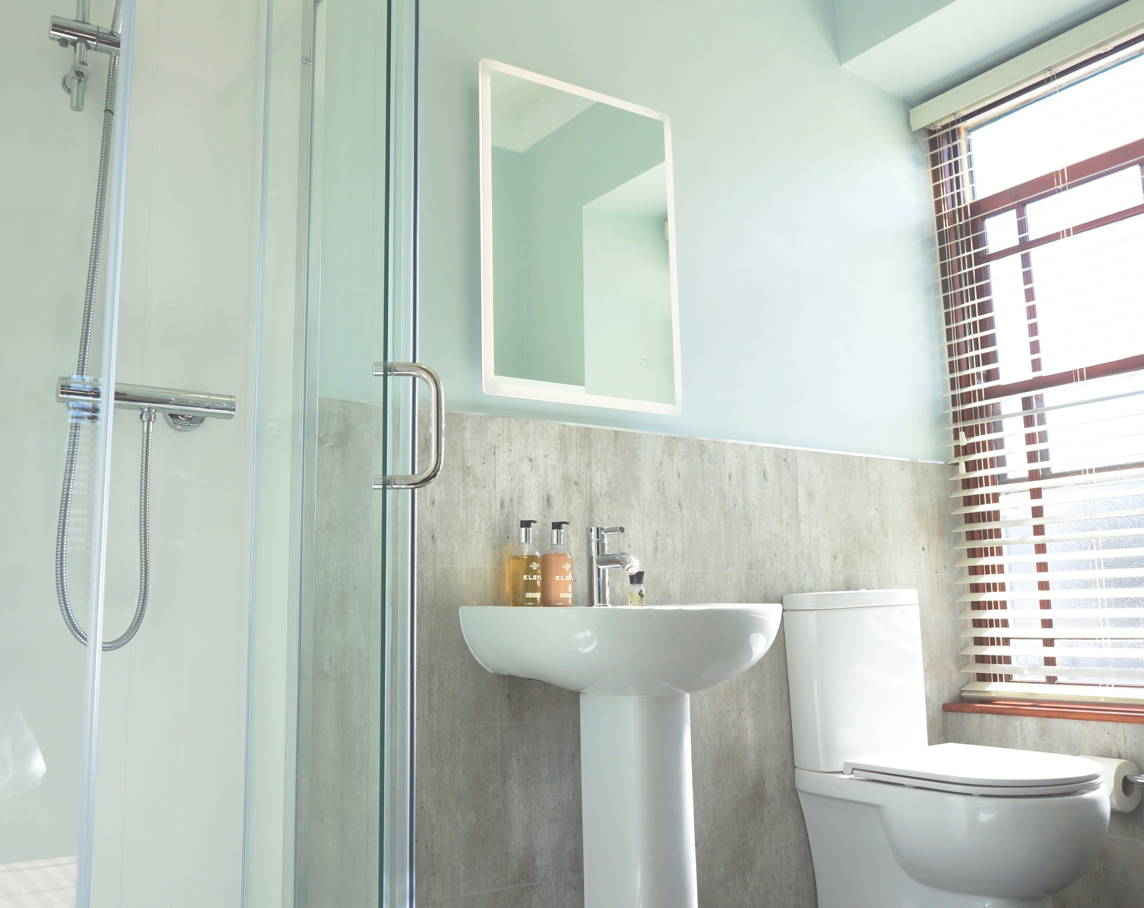 A bathroom with a glass shower enclosure, a white sink, a toilet, and a window with blinds.