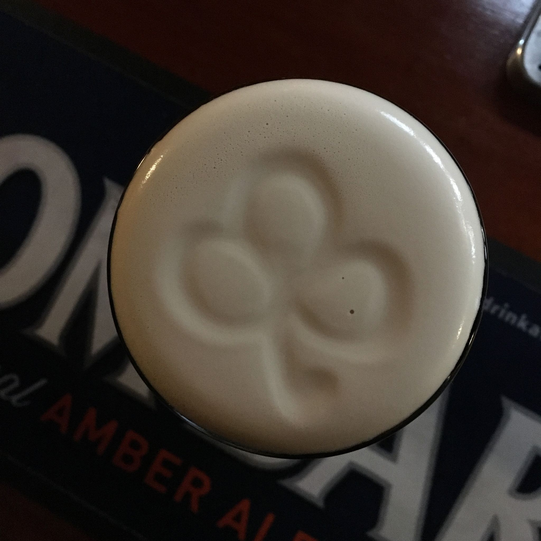 A cup of coffee with a frothy top, placed on a table with a coaster that has text partially visible.