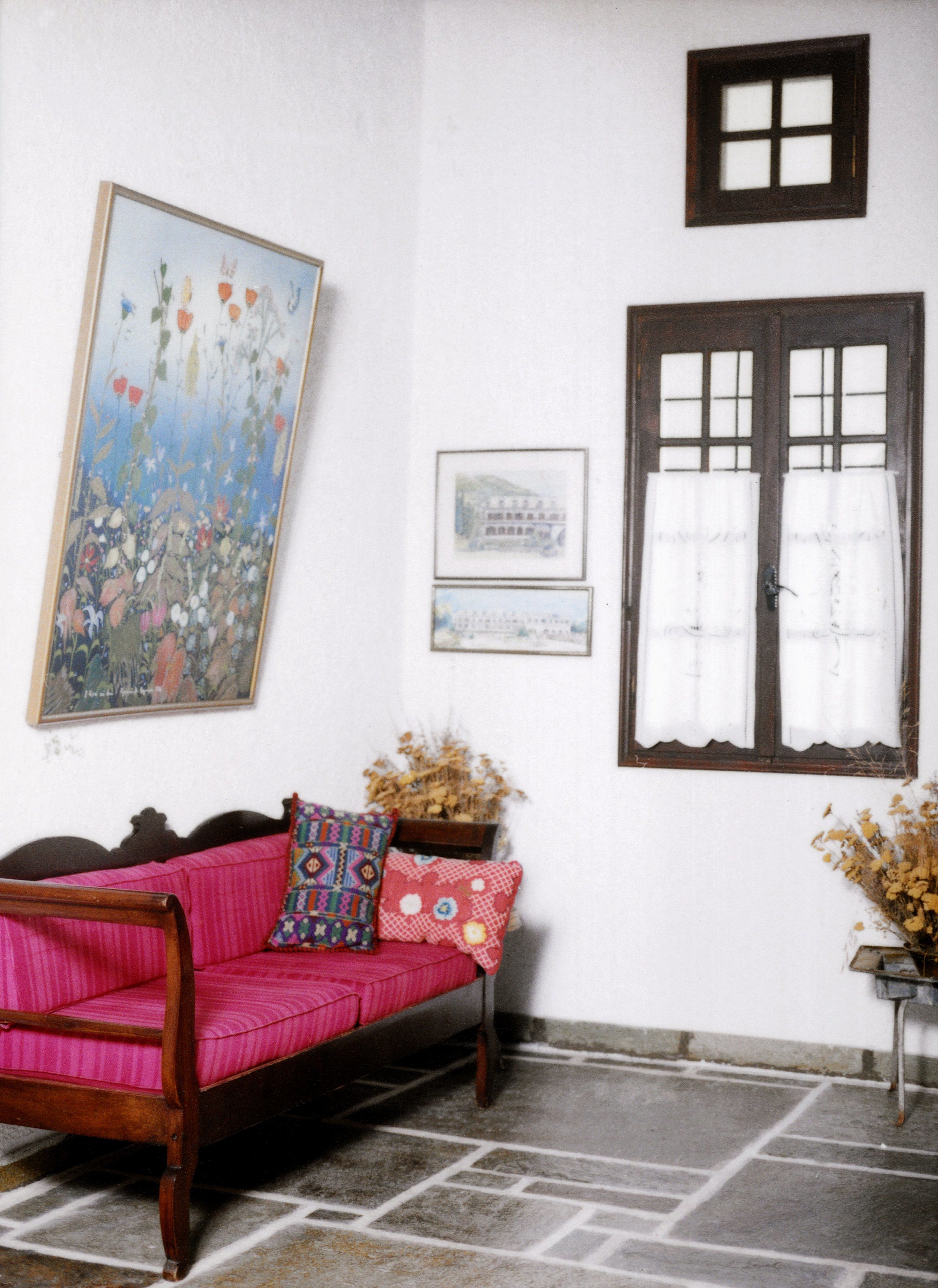 Una habitación con sofá de madera, un cuadro floral grande y una ventana con cortinas blancas.