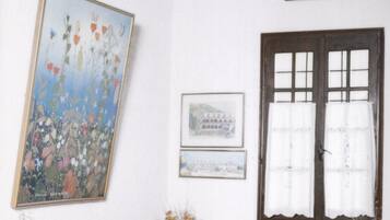 Una habitación con sofá de madera, un cuadro floral grande y una ventana con cortinas blancas.