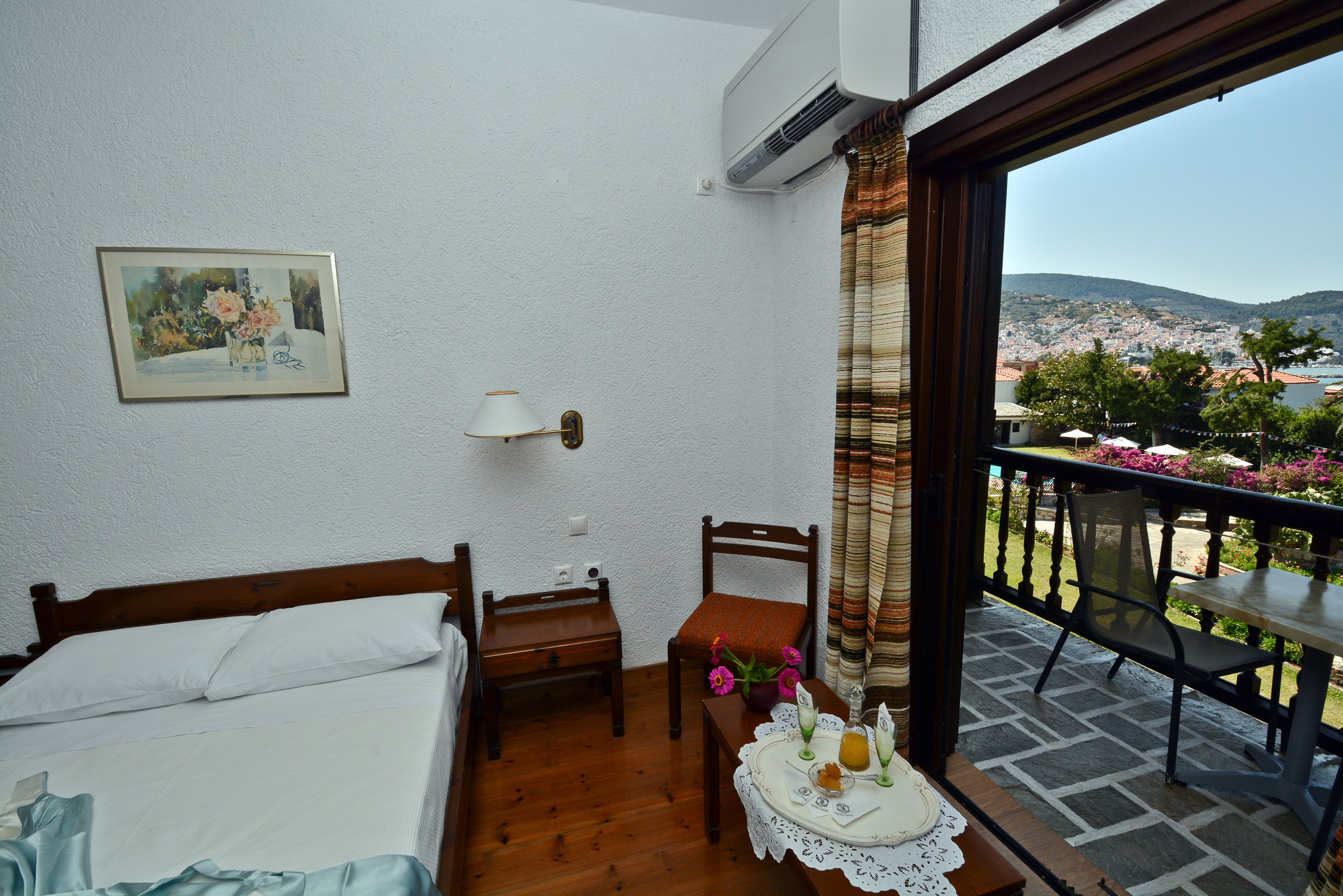 Quarto com cama, mesa de cabeceira, cadeira, mesa com um vaso de flores e vista para uma varanda com mesa e cadeiras.