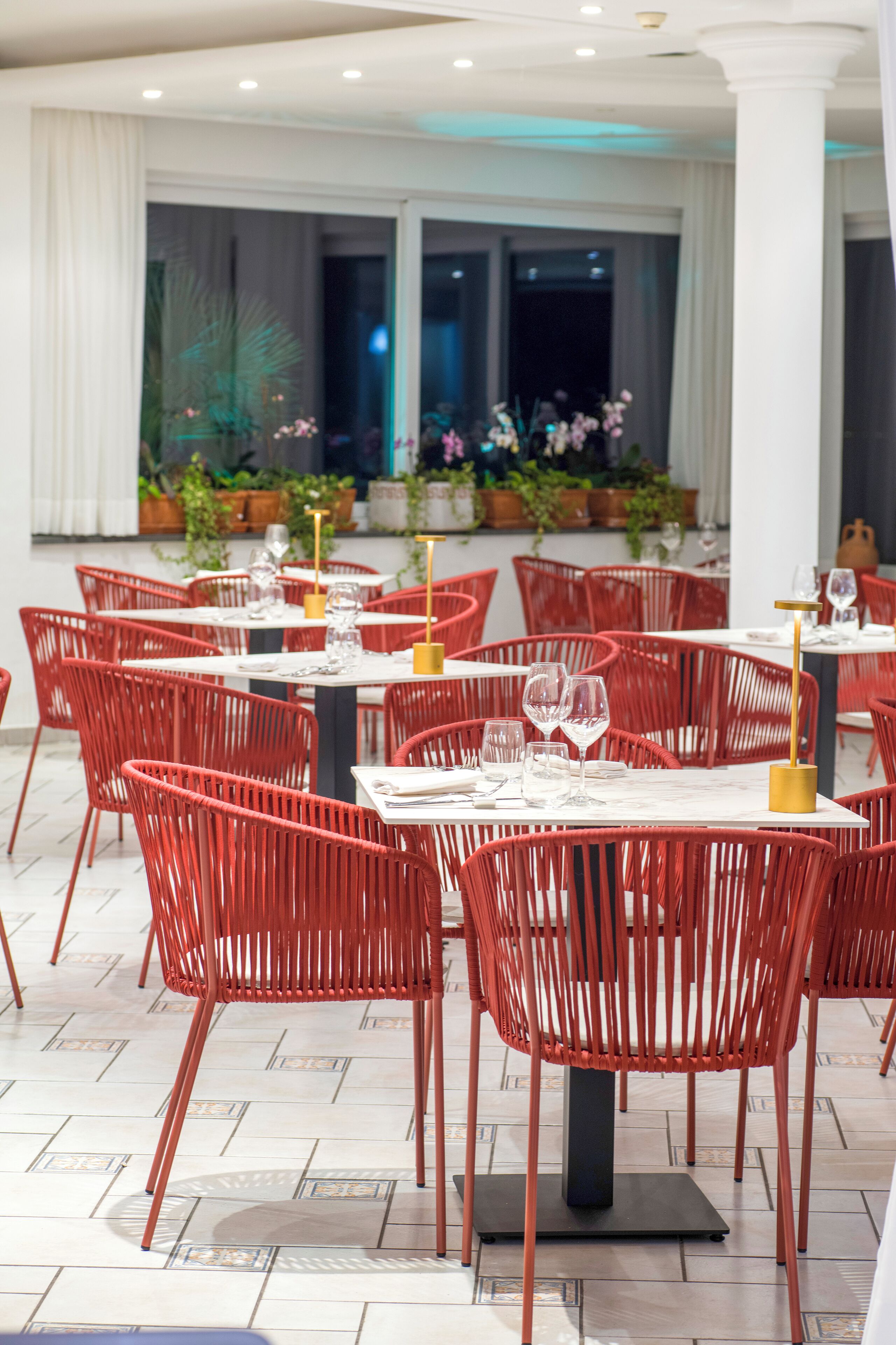 Ein Restaurant mit roten Stühlen und weißen Tischen, mit Fliesenboden und Pflanzen im Hintergrund.