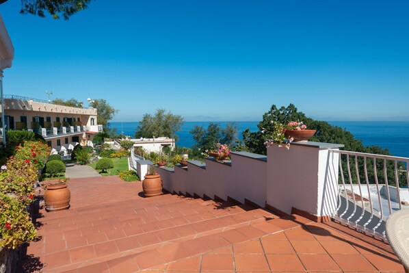 A terracotta-tiled outdoor area with potted plants, a white railing, and a view of the sea.