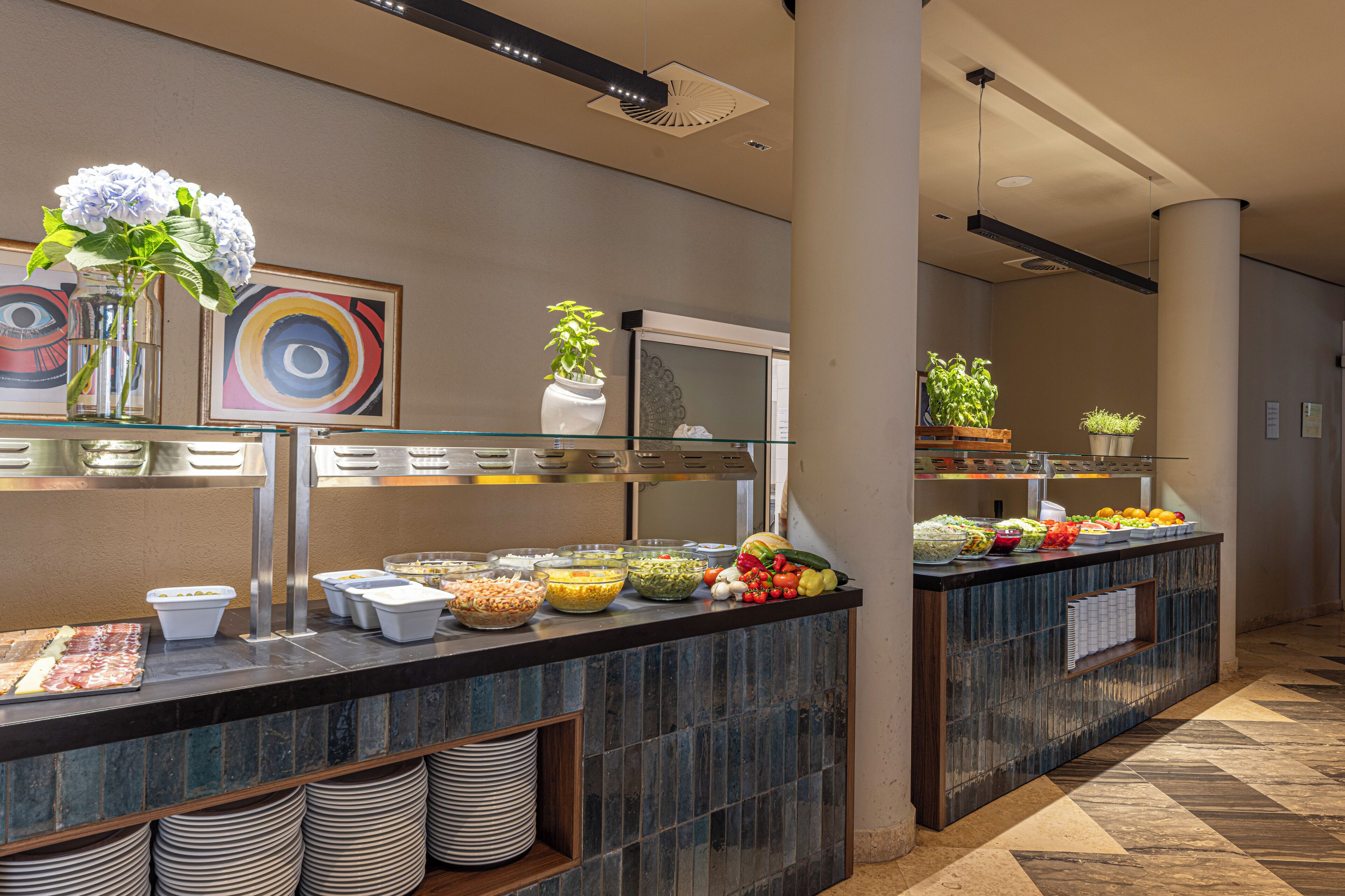 Un área de bufé con diversos alimentos, un expositor de vidrio y plantas decorativas.