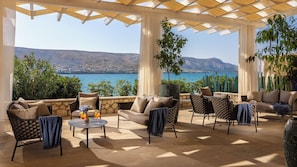 A covered outdoor seating area with stone flooring, cushioned seating, and a view of a lake and mountains. - Family Hotel Pagus (Pag)