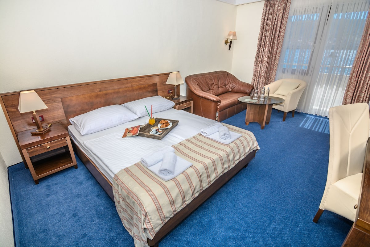 Une chambre d’hôtel avec un grand lit, deux tables de chevet, une tête de lit en bois, un fauteuil en cuir marron, un fauteuil blanc, une petite table ronde et une fenêtre avec des rideaux légers.
