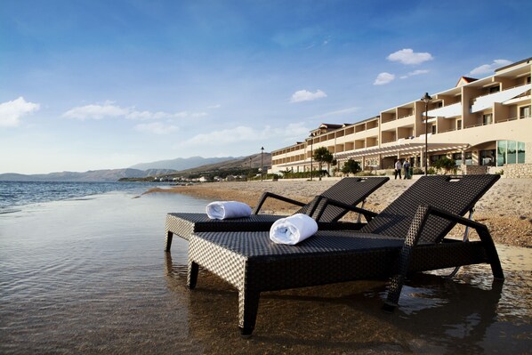 A beachside hotel with lounge chairs and towels on the sand, overlooking the sea and mountains. - Family Hotel Pagus (Pag)