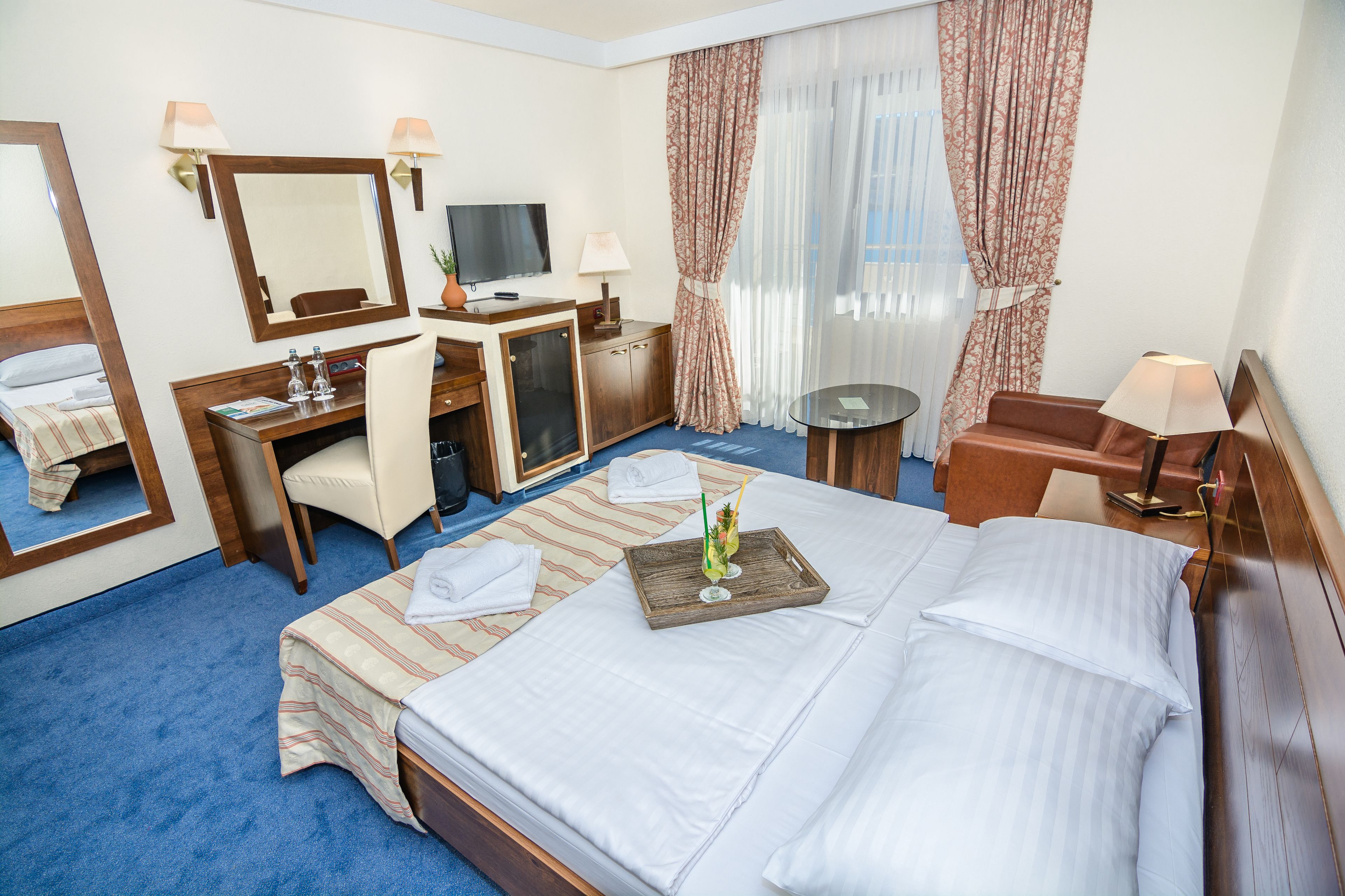 A hotel room with a bed, a desk, a chair, a TV, and a small table with a tray.