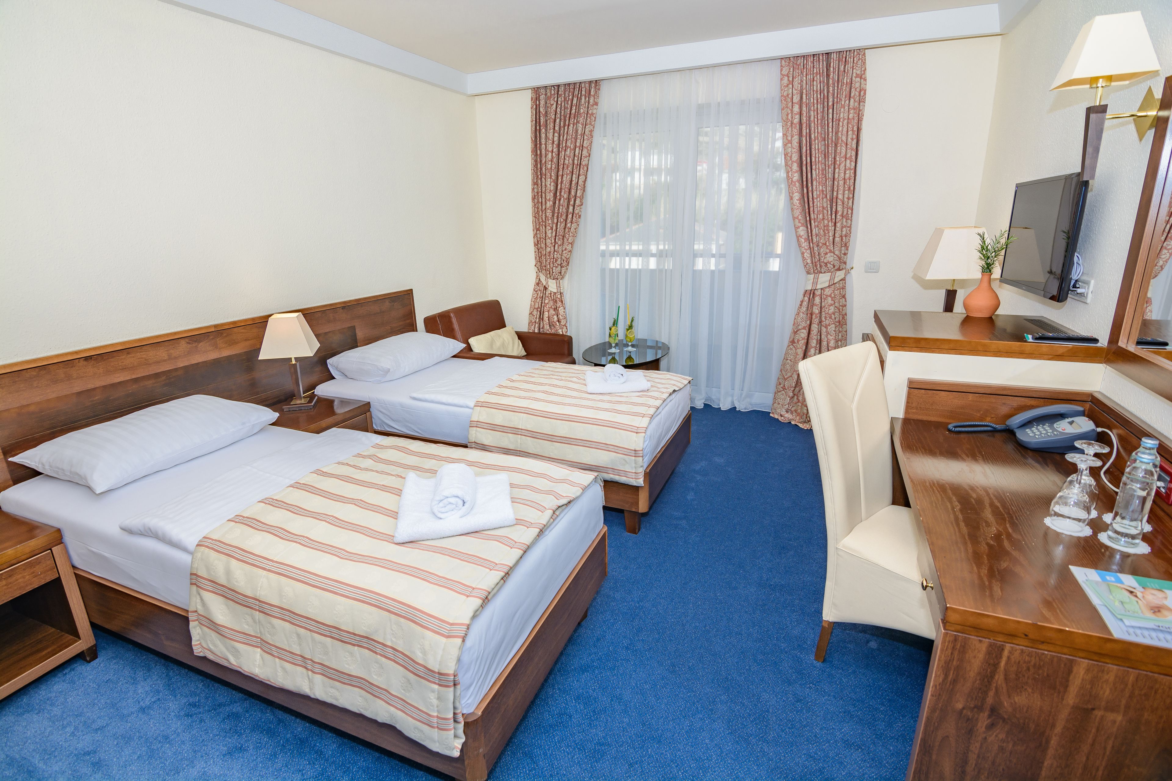 A hotel room with two beds, a desk with a telephone, a TV, and a small table with a lamp and decorative items.