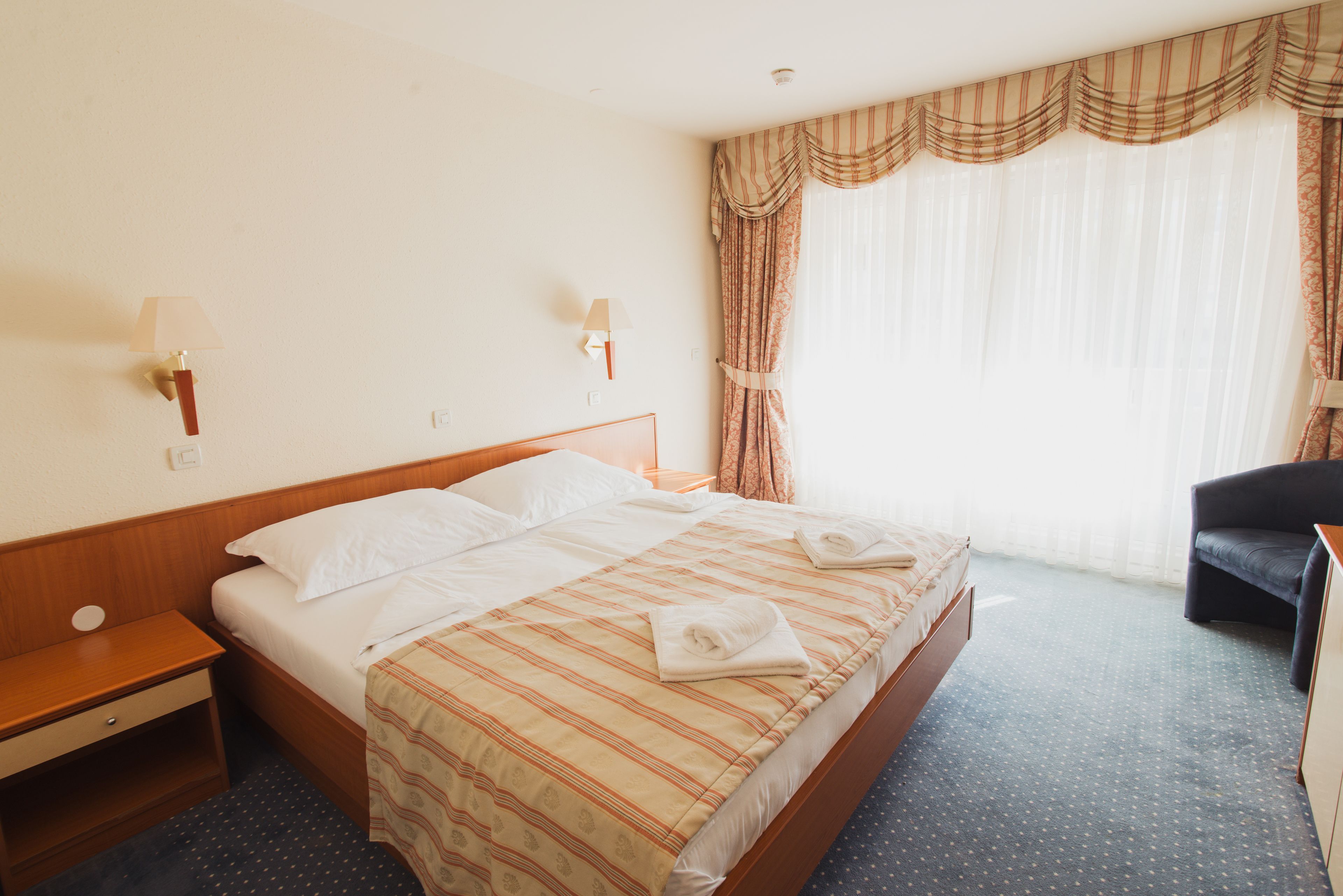 Een hotelkamer met een bed, twee nachtkastjes, een stoel en een raam met gordijnen.