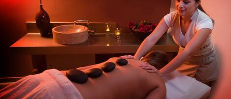 Eine Massageanwendung in einer Wellnessumgebung mit einer Massageliege, einer Schale mit heißen Steinen und einer Blumenvase.