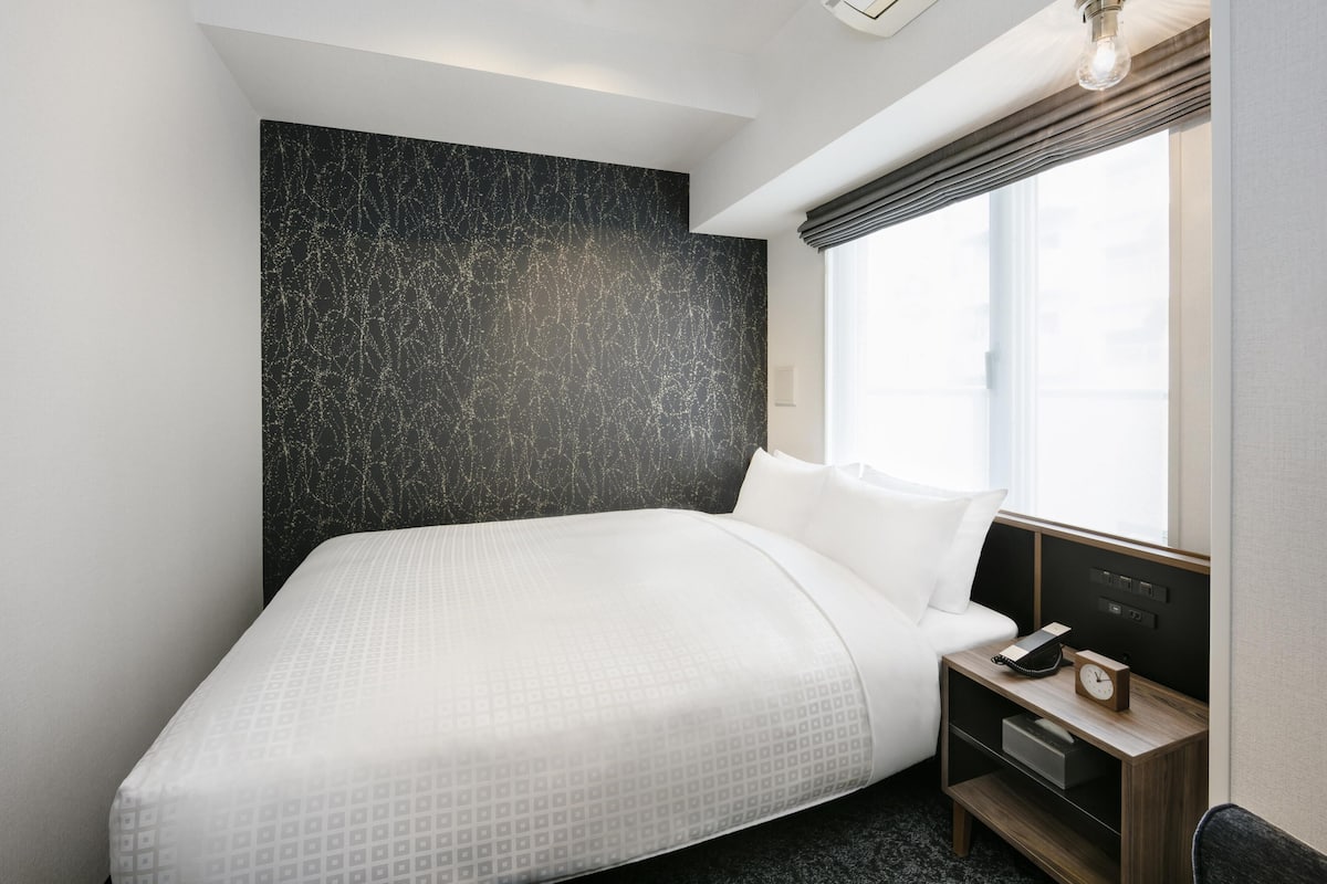 Een moderne hotelkamer met een groot bed, een nachtkastje met telefoon en wekker, een raam met gordijnen en een wandairconditioning.