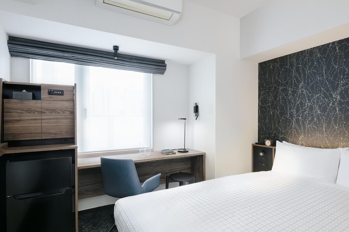 Hotelkamer met een bed, een bureau, een stoel, een kast, een nachtkastje, een lamp en een wandklok.
