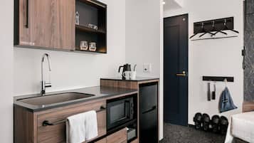 A compact hotel room with a kitchenette, a dark door, a coat rack, and a bed with a patterned bedspread.