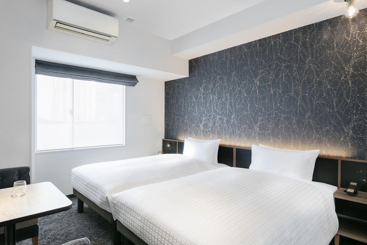 Een hotelkamer met twee bedden, een tafeltje, een stoel en een groot raam.