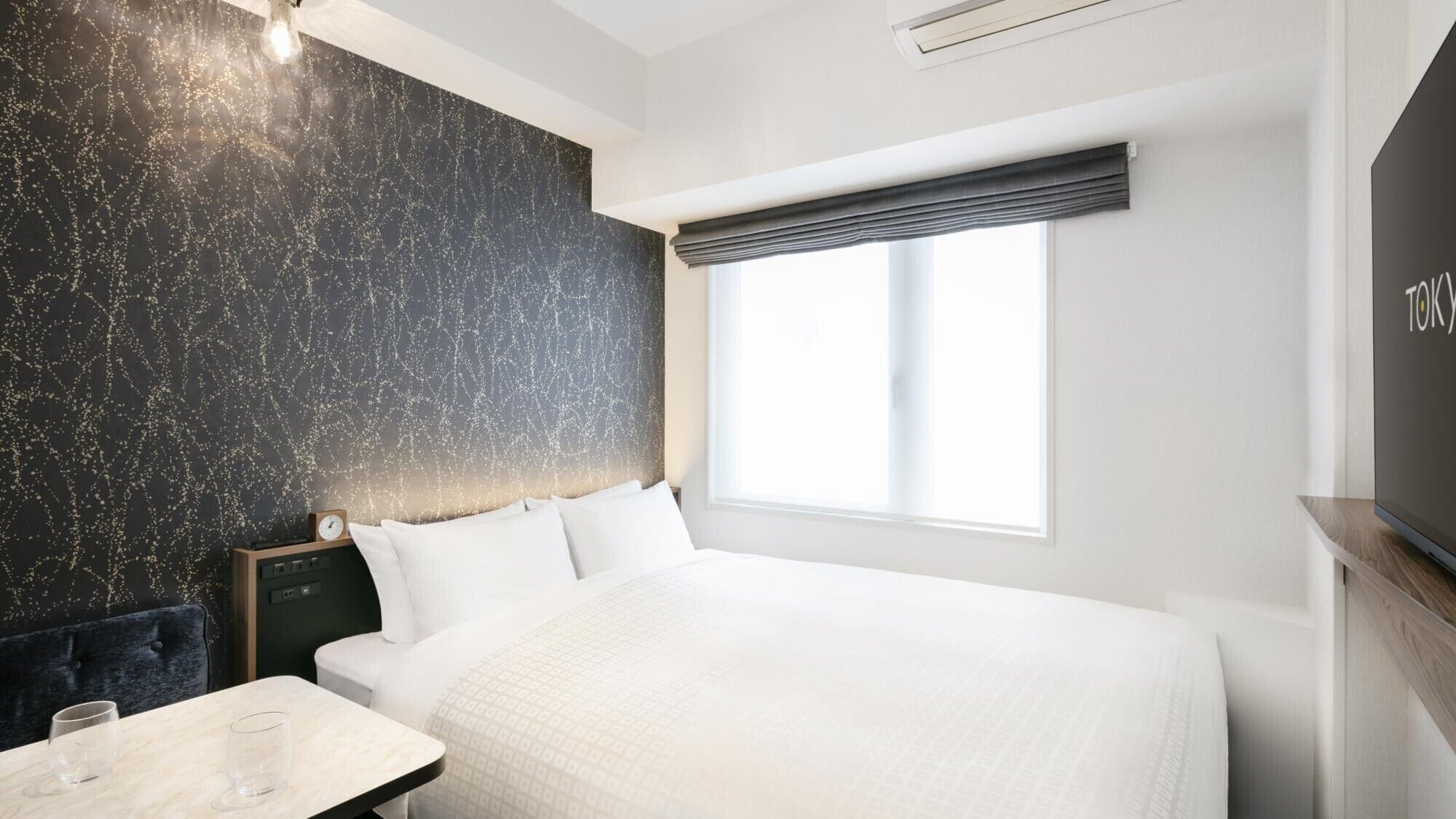 Una camera d'albergo moderna con un letto grande, un comodino con bicchieri, una sveglia e una finestra vista.