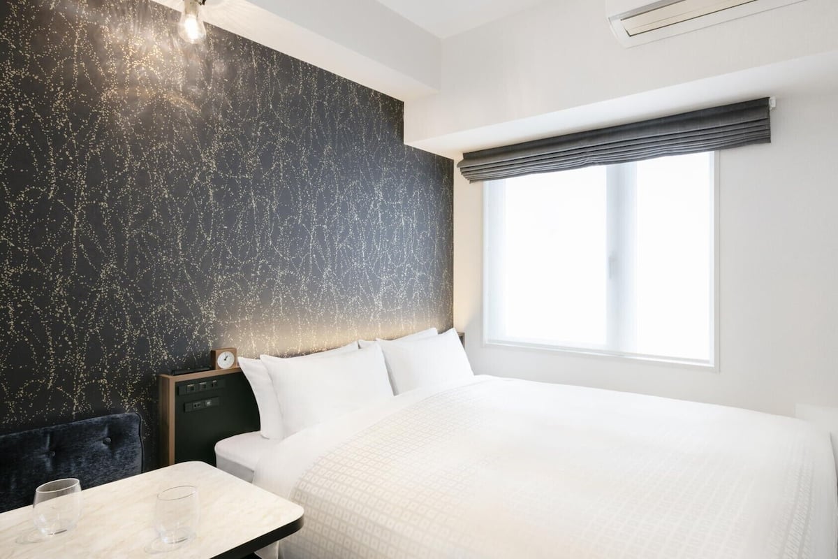 Een moderne hotelkamer met een groot bed, een nachtkastje met glazen, een klok en een raam met uitzicht.