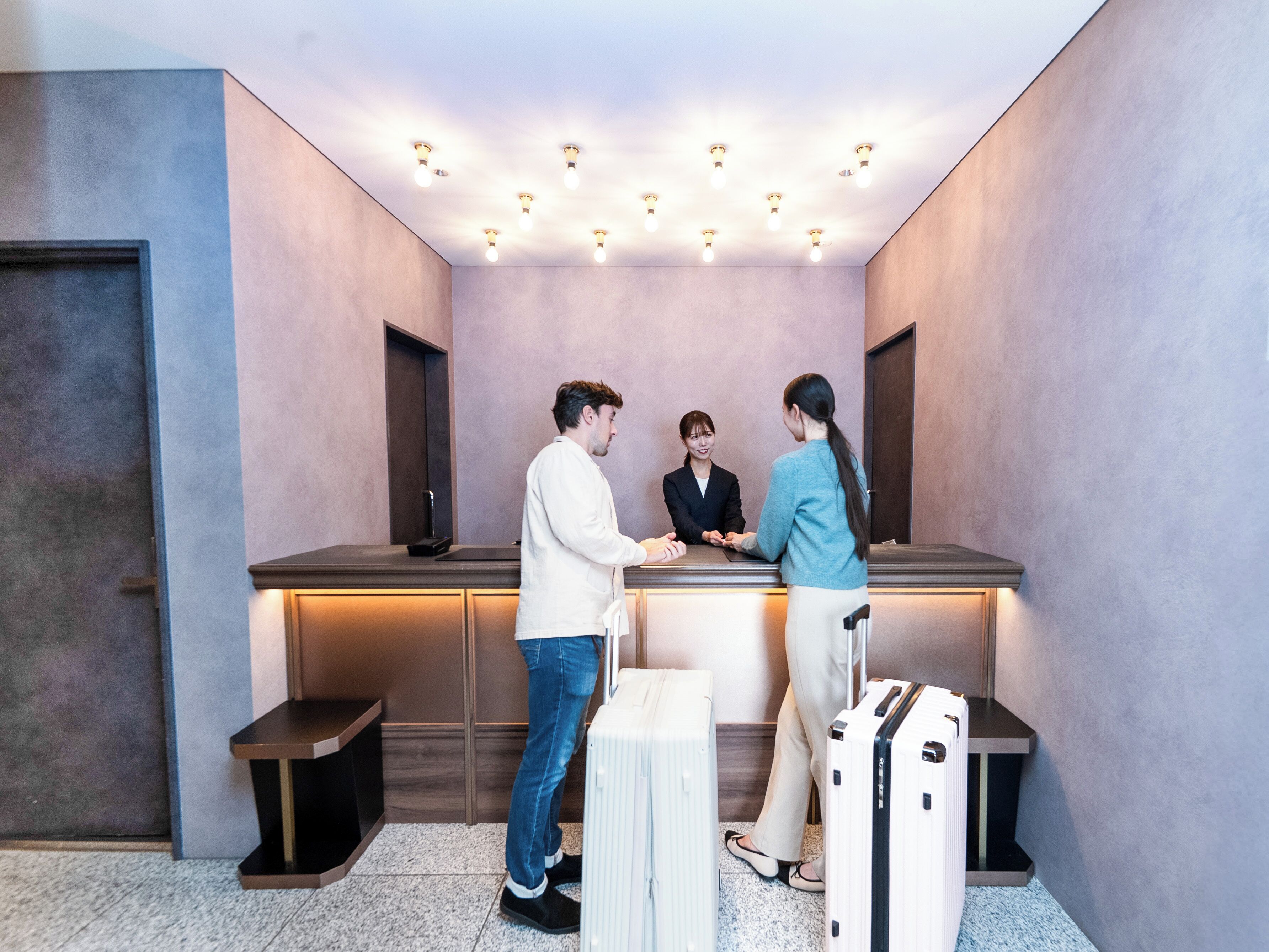 A reception area with a reception desk, a counter, and a seating bench. There are two suitcases and a handbag on the floor.