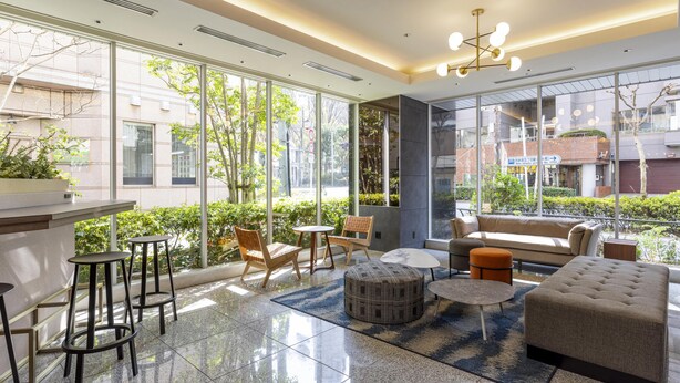 Un espace de détente moderne comprenant un canapé, un pouf, des fauteuils et une petite table. De grandes fenêtres offrent une vue sur l’extérieur.