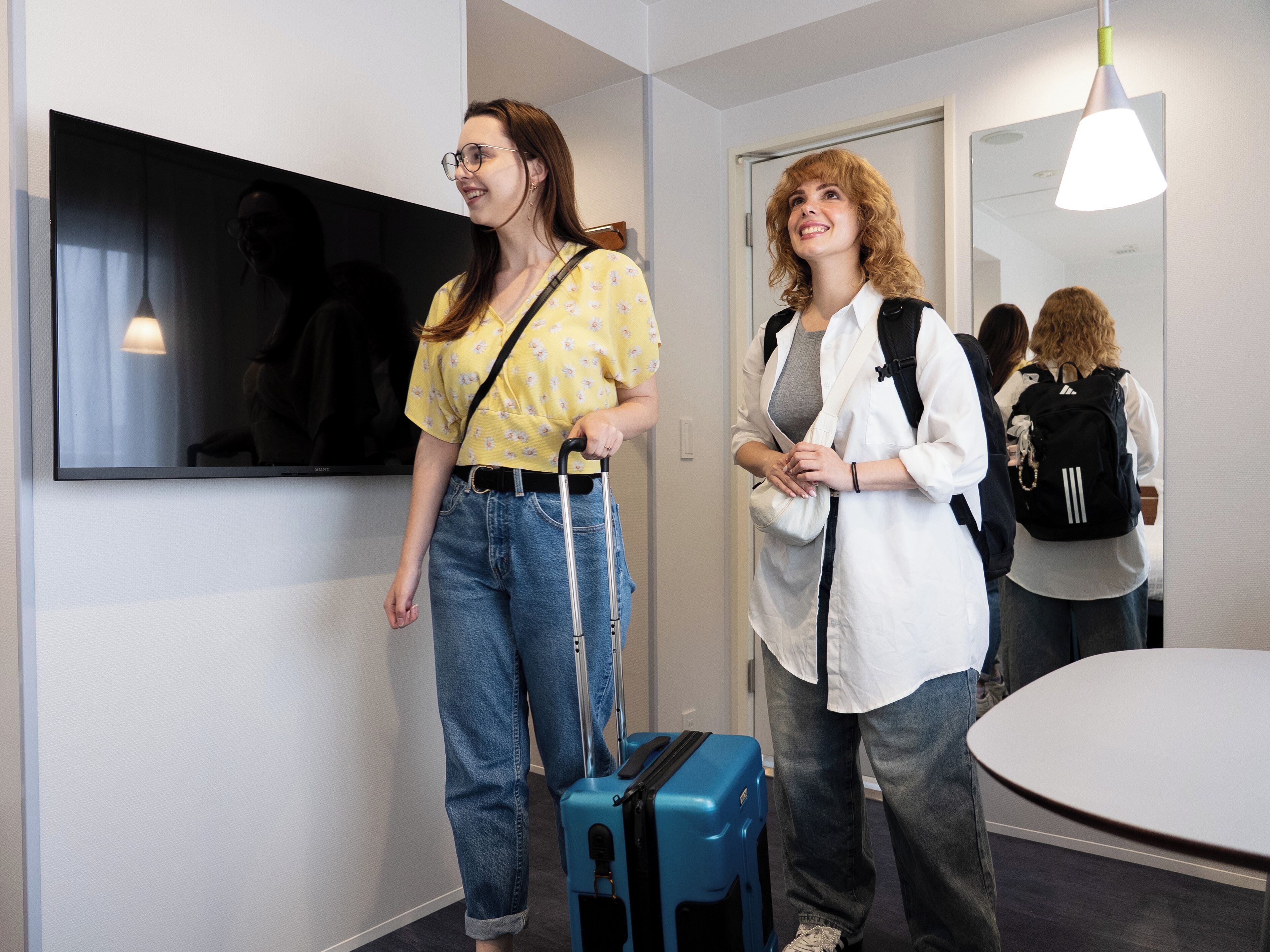 A hotel room with a TV mounted on the wall, a round table, and a blue suitcase.
