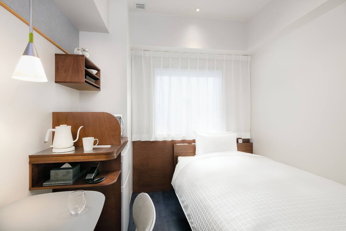 A hotel room with a bed, a desk with a kettle, a chair, and a window with curtains.