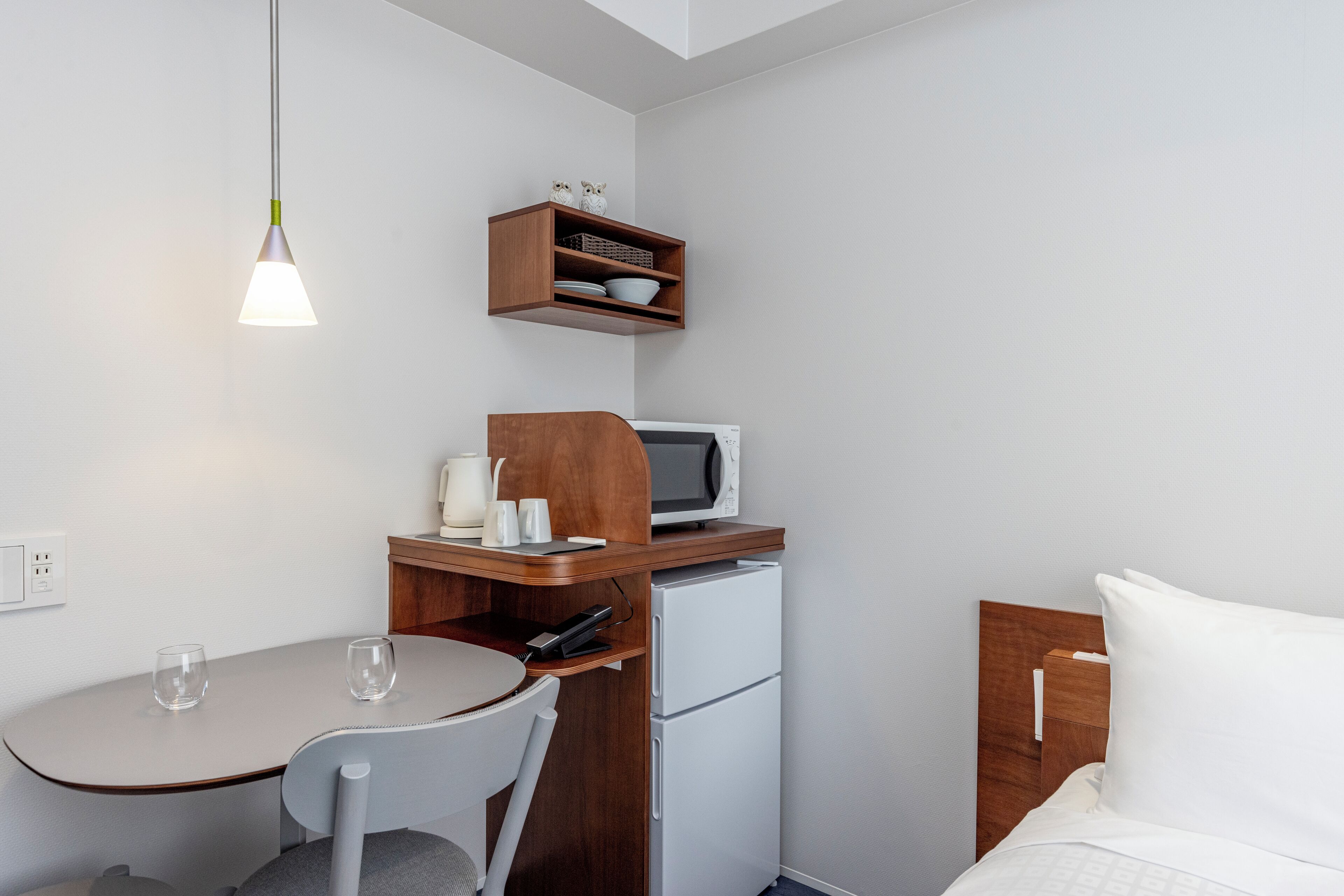 A compact kitchenette with a microwave, a small refrigerator, a wooden shelf with kitchenware, a table with two chairs, and a hanging light fixture.