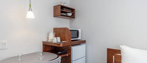 A compact kitchenette with a microwave, a small refrigerator, a wooden shelf with kitchenware, a table with two chairs, and a hanging light fixture.
