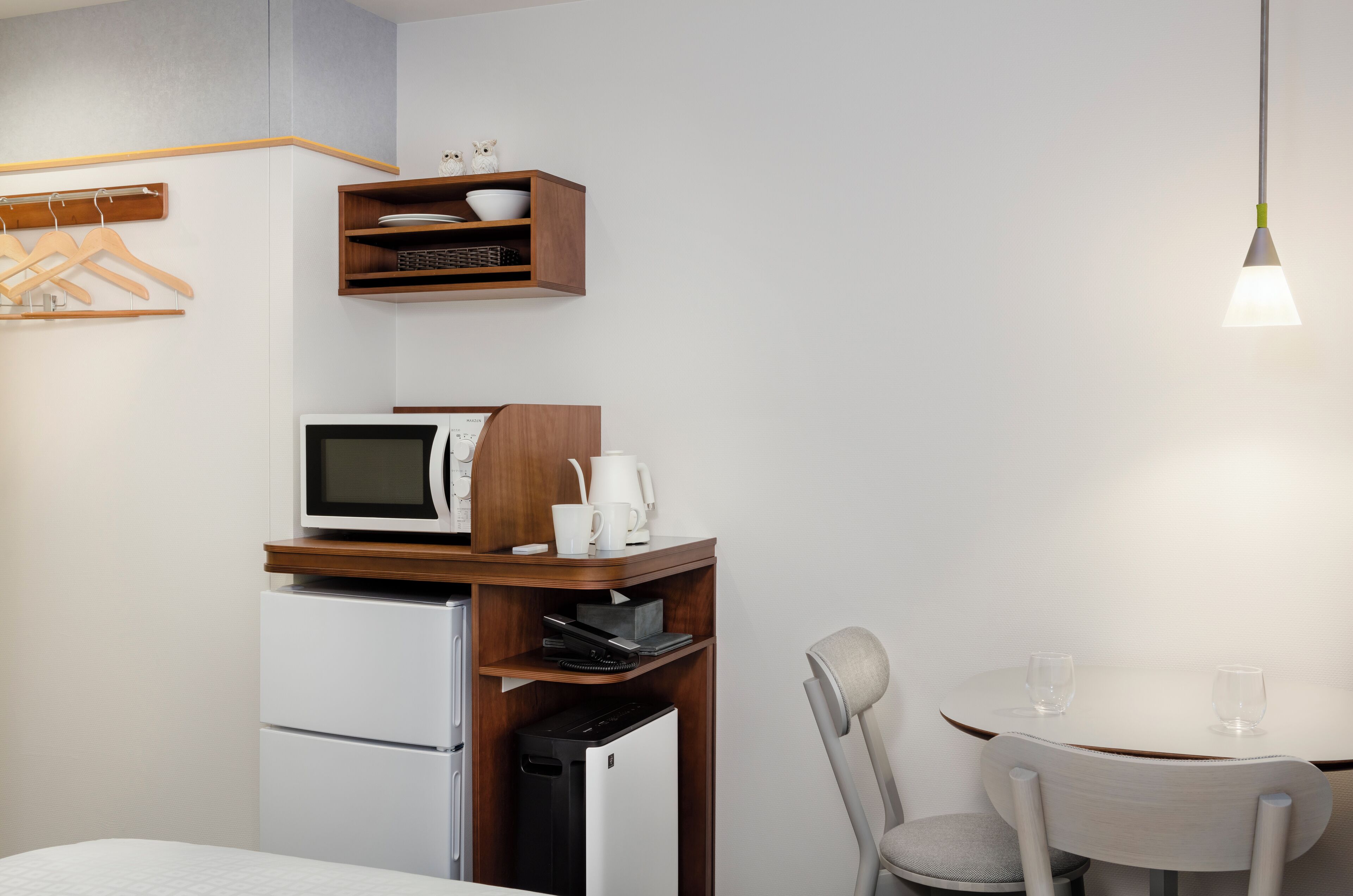 Uma cozinha compacta equipada com micro-ondas, cafeteira e pequena geladeira. Há uma prateleira de madeira acima do micro-ondas, e uma mesa com duas cadeiras.