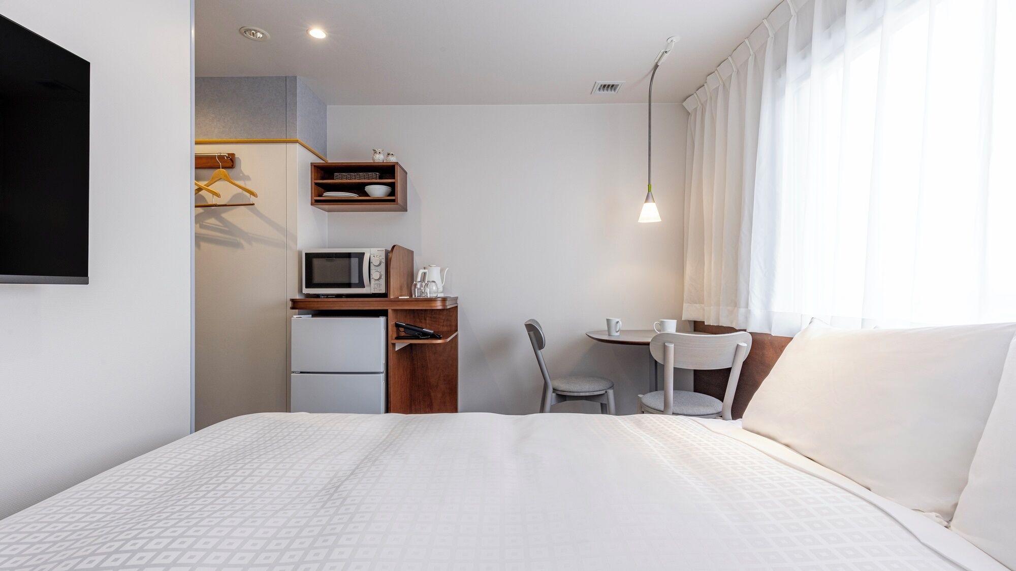 Habitació d'hotel amb un llit, escriptori amb microones, nevera, una cadira, taula petita i televisió penjada a la paret.