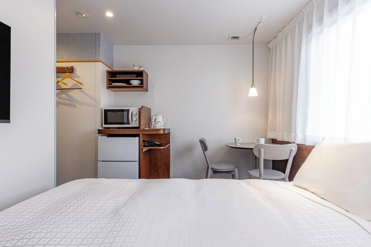 A hotel room with a bed, a desk with a microwave, a refrigerator, a chair, a small table, and a TV mounted on the wall.