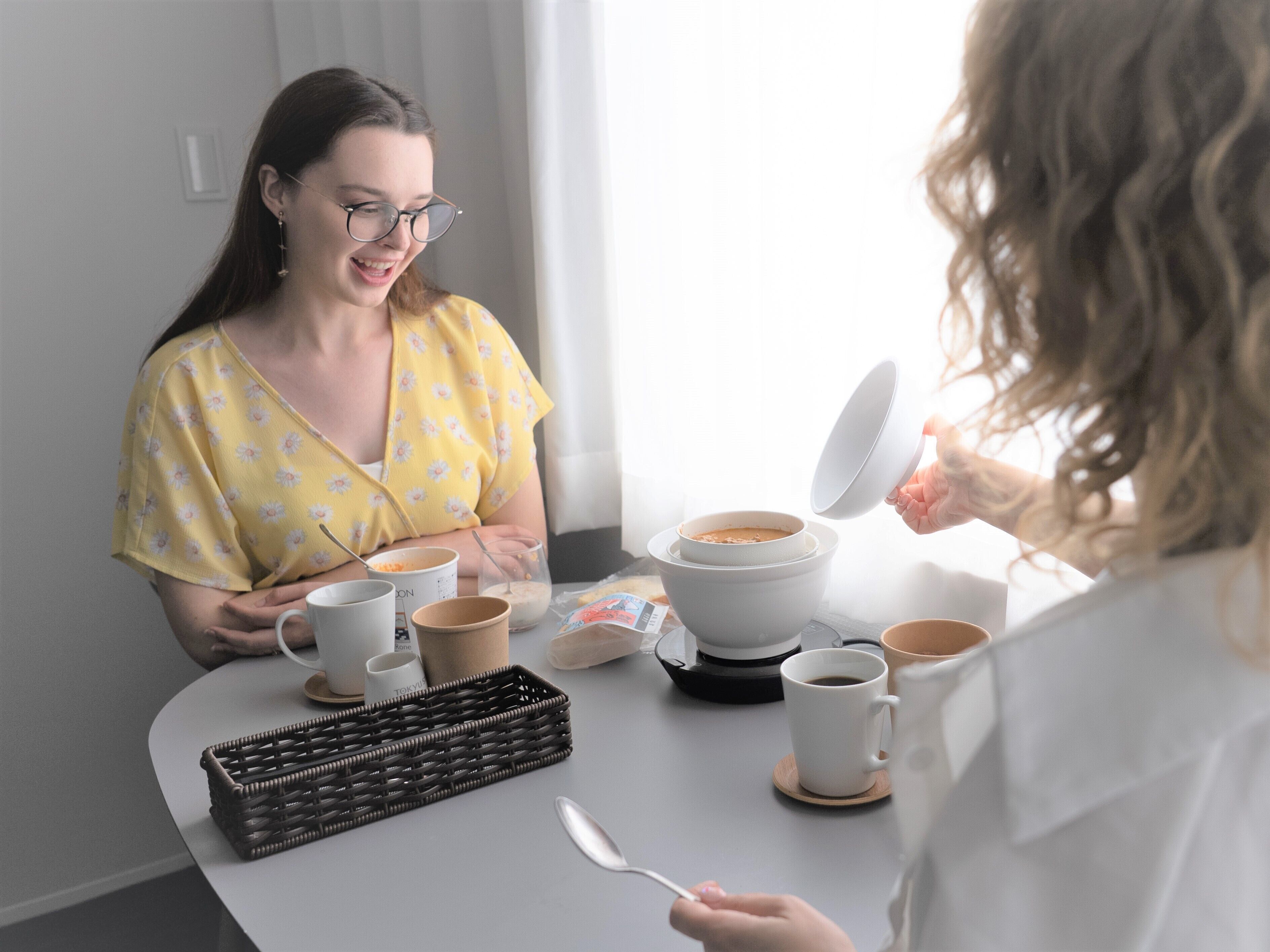 A breakfast setting with a coffee maker, cups, and a basket on a table.