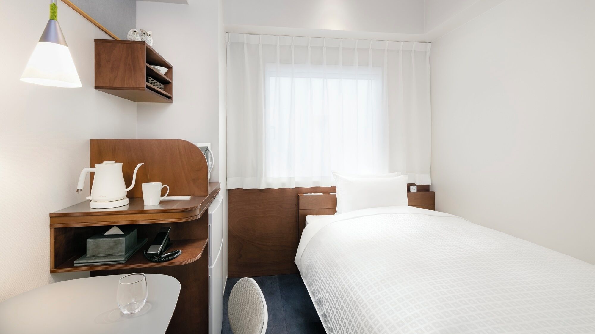 Habitació d'hotel amb un llit, una taula de treball amb una cafetera, una cadira, una estantària amb objectes decoratius i una finestra amb cortines primes.