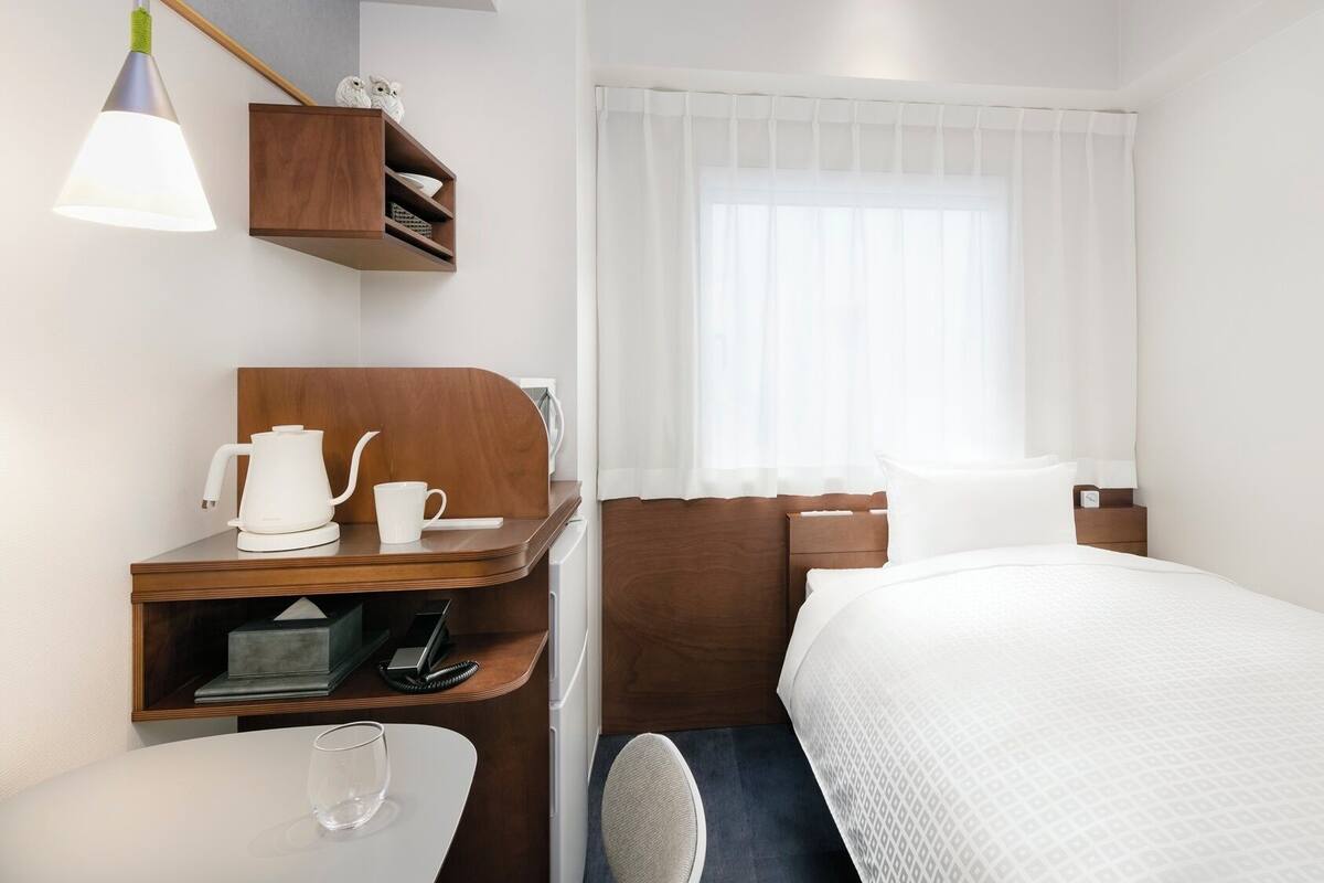 A hotel room with a bed, a desk with a coffee maker, a chair, a shelf with decorative items, and a window with sheer curtains.