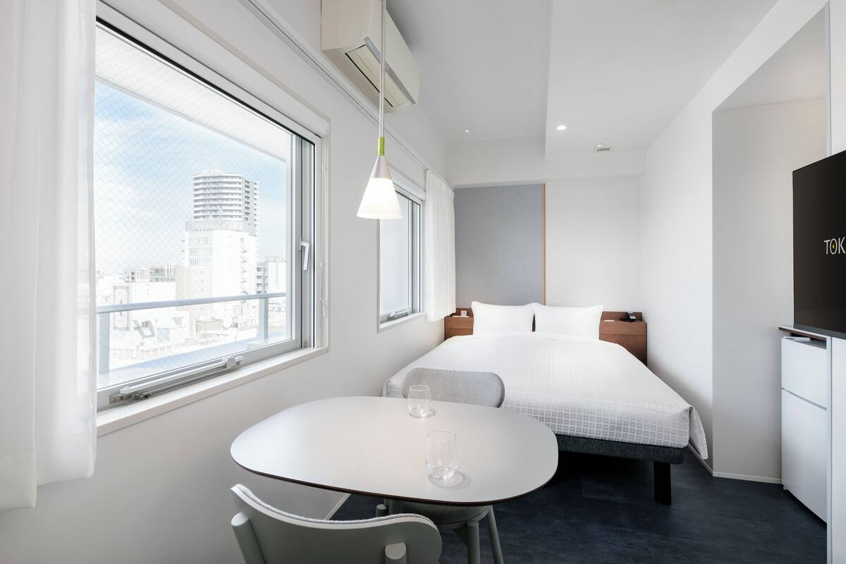 A modern hotel room with a bed, bedside table, and a window with a city view.